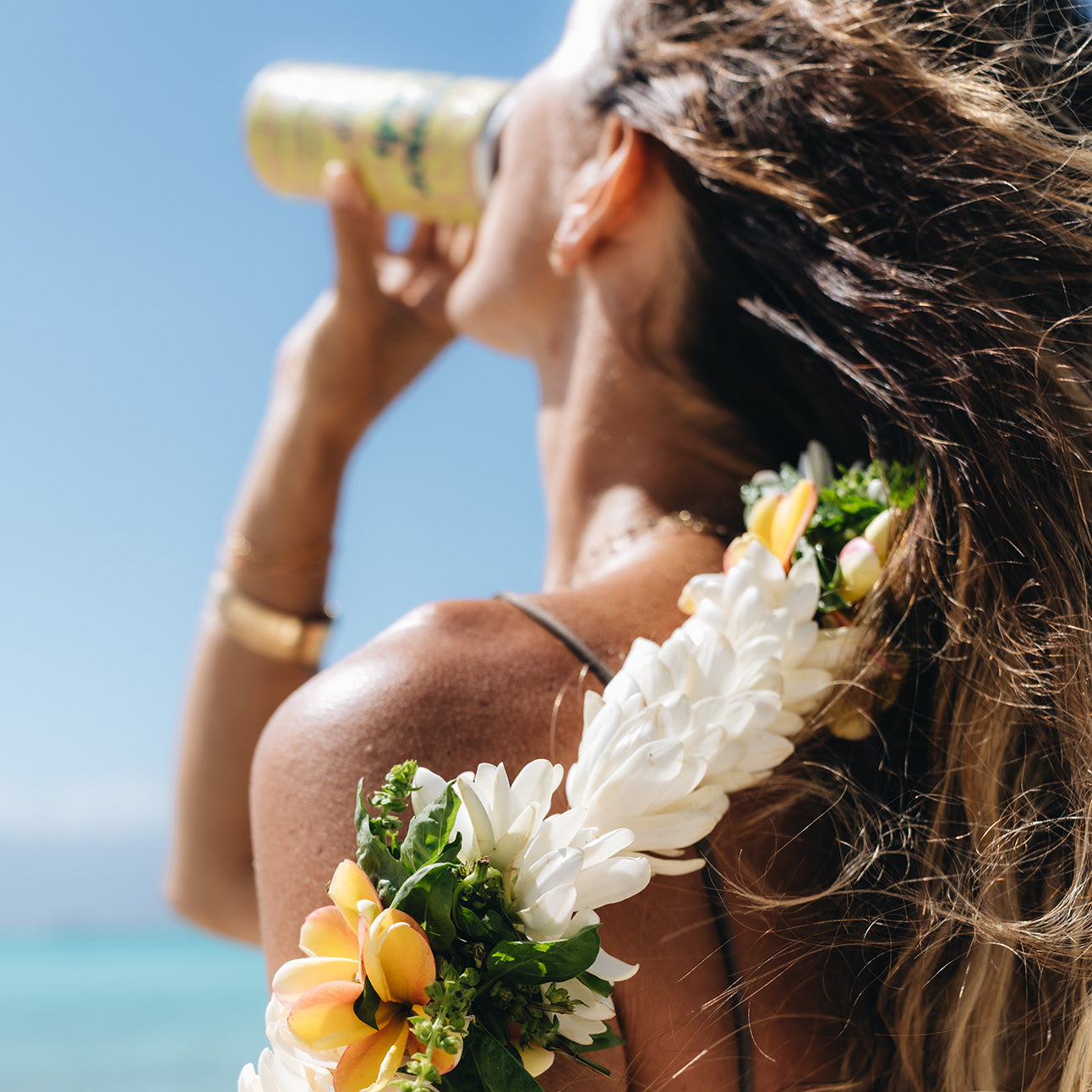 Woman with a floral wreath on her head drinking Tahitian Spritz against a blue sky.